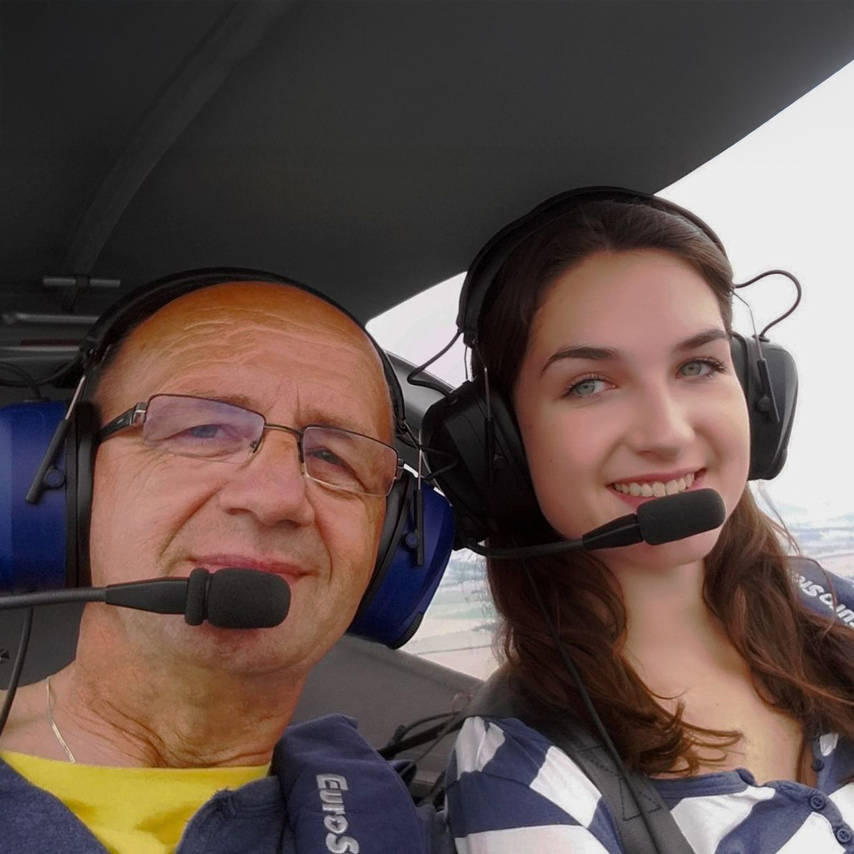 Zwei Personen mit Headsets im Cockpit eines Ultraleichtflugzeugs, lächelnd in die Kamera.