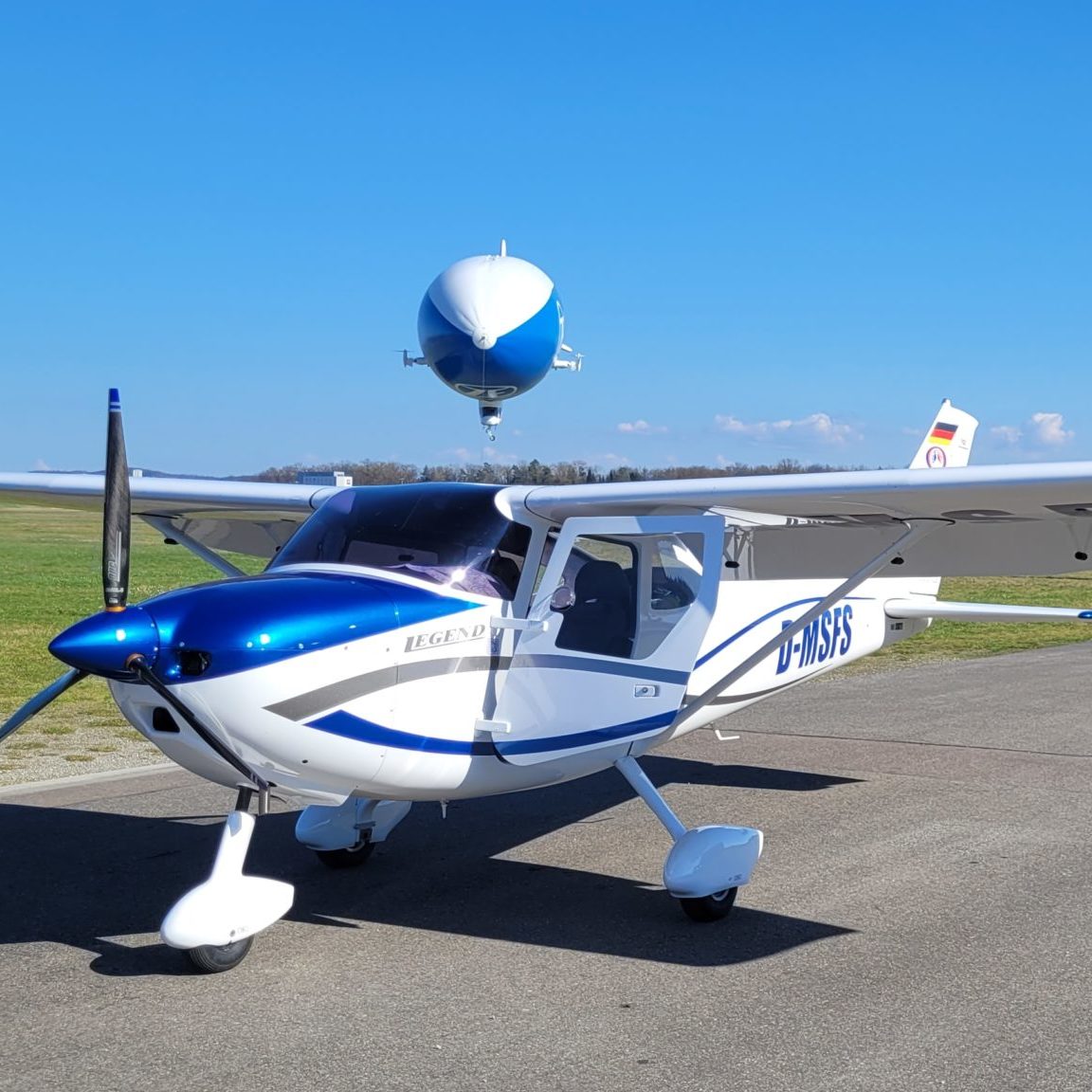 Leichtflugzeug mit der Kennung D-MSFS auf dem Rollfeld, im Hintergrund ein überfliegender Zeppelin am klaren, blauen Himmel.