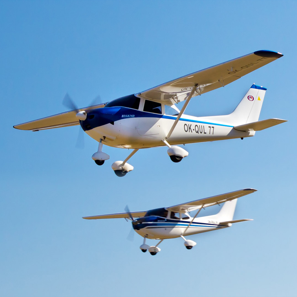 Zwei weiß-blaue Leichtflugzeuge des Typs Legend fliegen in Formation am klaren, blauen Himmel.