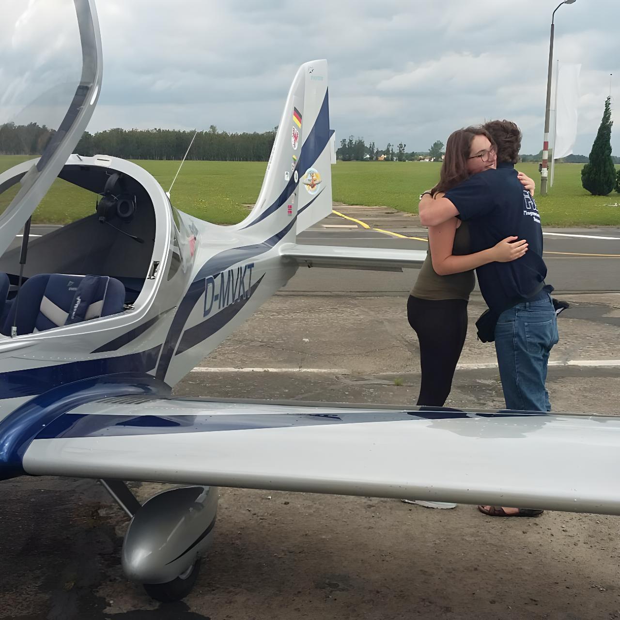Zwei Personen umarmen sich herzlich neben einem abgestellten Leichtflugzeug mit offener Cockpithaube auf dem Flugplatz.