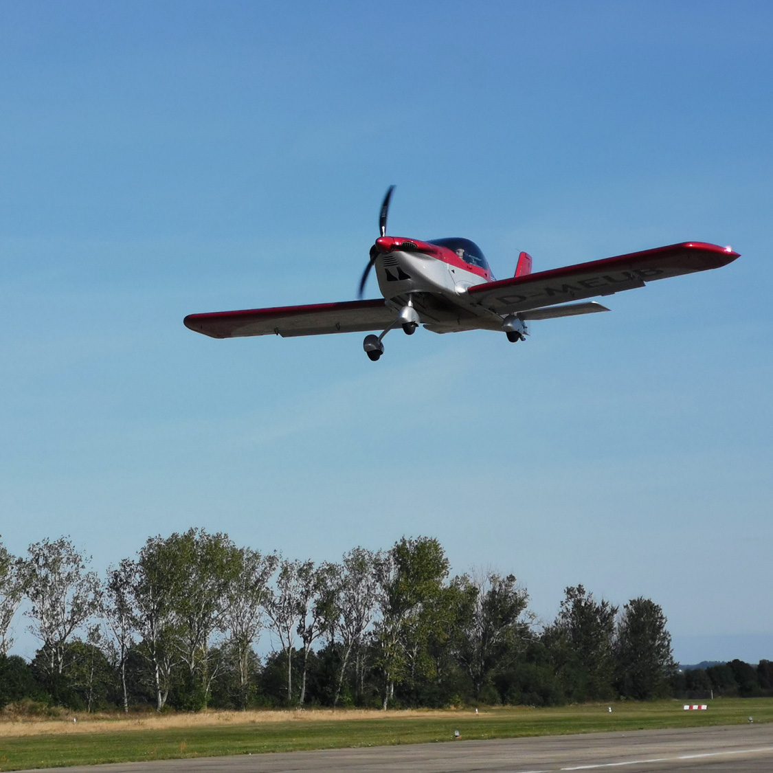 Leichtflugzeug hebt bei blauem Himmel von der Startbahn ab, im Hintergrund eine Baumreihe am Rand des Flugplatzes.