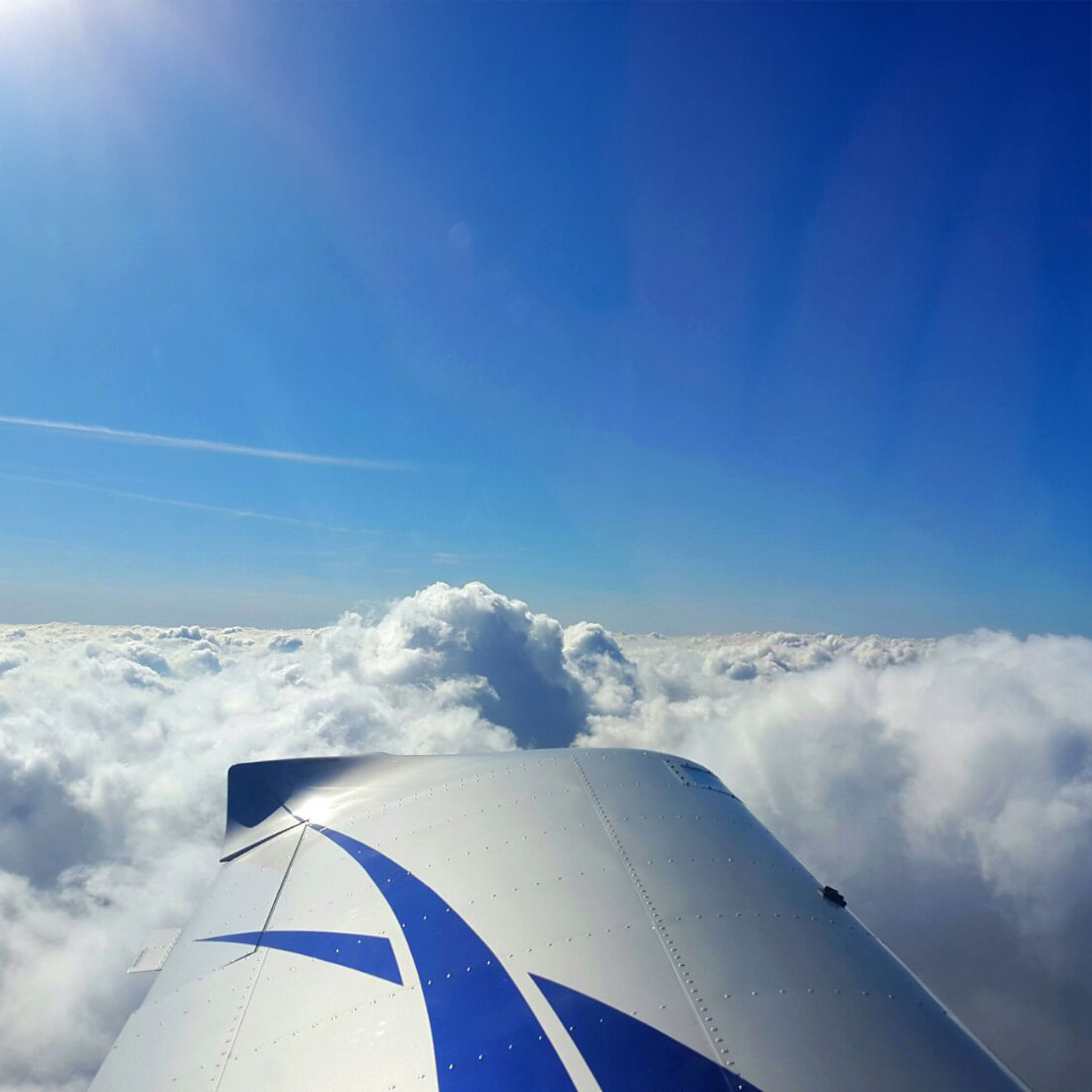 Blick aus dem Flugzeug über die Tragfläche hinweg auf eine geschlossene Wolkendecke unter strahlend blauem Himmel.
