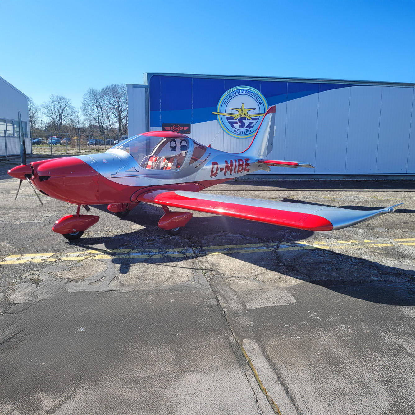 Leichtflugzeug in Rot und Silber mit der Kennung D-MIBE auf dem Vorfeld vor einer Halle des Flugplatzes Bautzen.