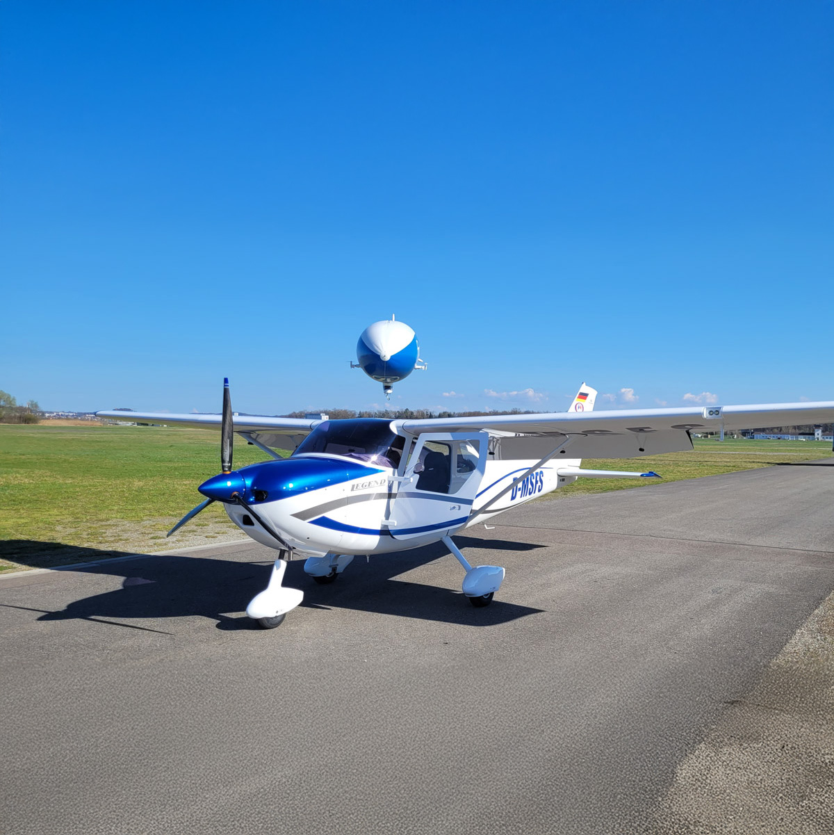 Leichtflugzeug mit der Kennung D-MSFS auf dem Rollfeld, im Hintergrund ein überfliegender Zeppelin am klaren, blauen Himmel.