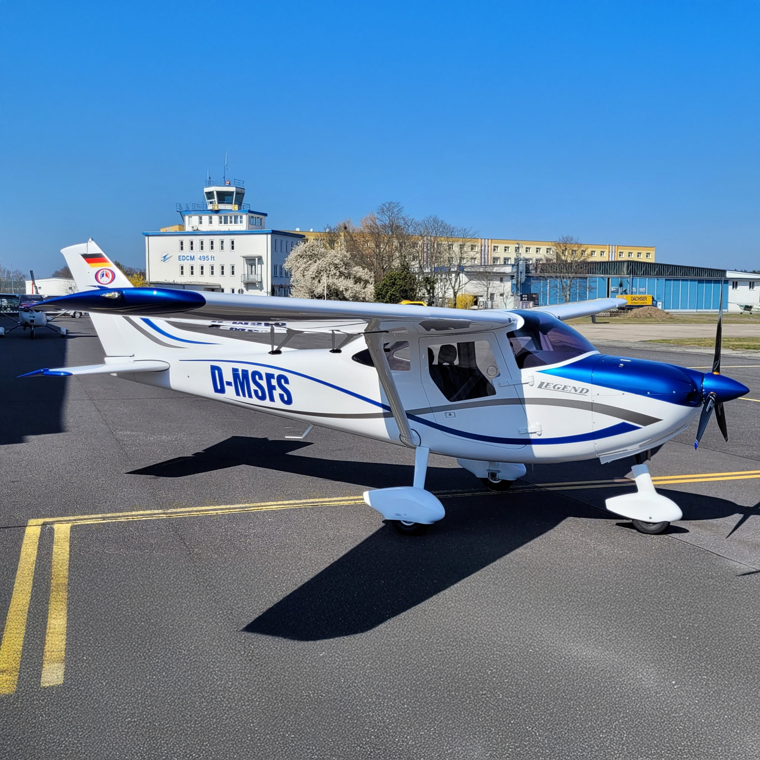 Kleinflugzeug mit der Kennung D-MSFS auf einem Flugplatz vor einem Towergebäude bei klarem, blauem Himmel.
