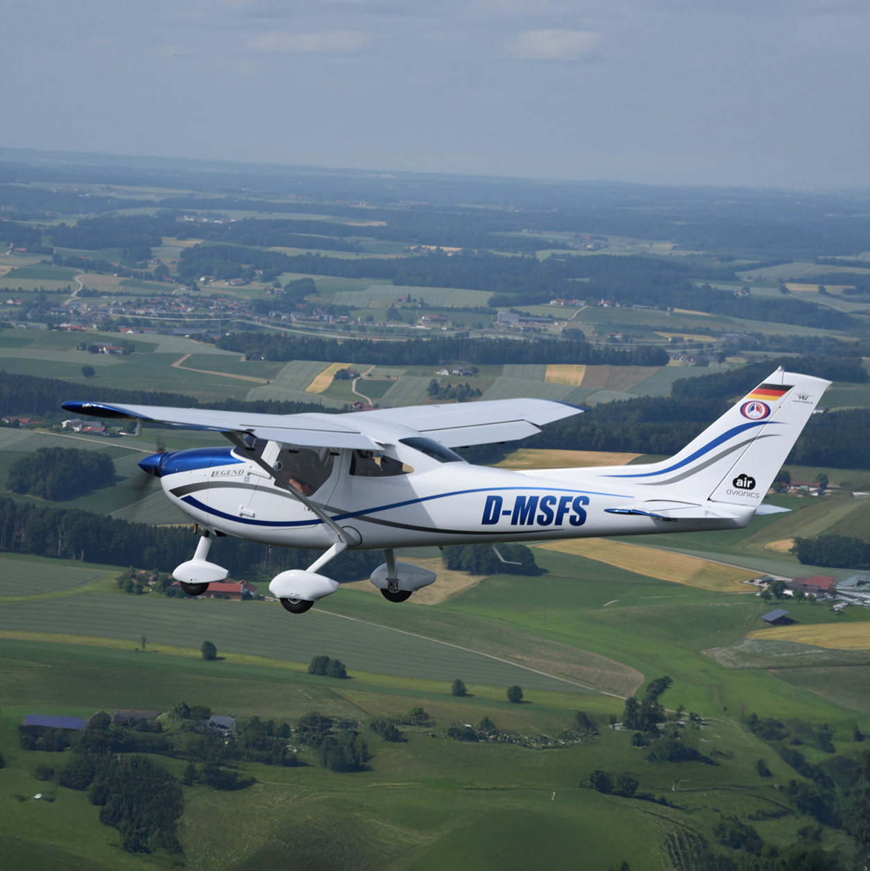 Weiß-blaues Leichtflugzeug mit der Kennung D-MSFS im Flug über einer ländlichen Landschaft mit Feldern und Wäldern.